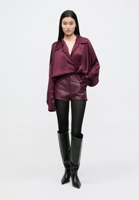 Bordo satino oversize marškiniai, odinės šortai ir juodi kelių aukščio aulinukai ant aukšto stiletto kulno. Lygi tekstūra, smailus galiukas, aptempta siluetas.