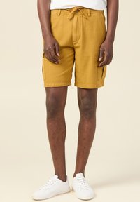 Bermudas cargo amarillas mostaza con un tejido texturizado, que cuentan con una cintura elástica con cordón y bolsillos laterales. Combinadas con zapatillas blancas.