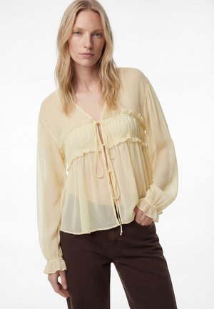 Mujer rubia con blusa translúcida de manga larga en amarillo pálido, con volantes y lazos, combinada con pantalones marrón oscuro sobre un fondo blanco.