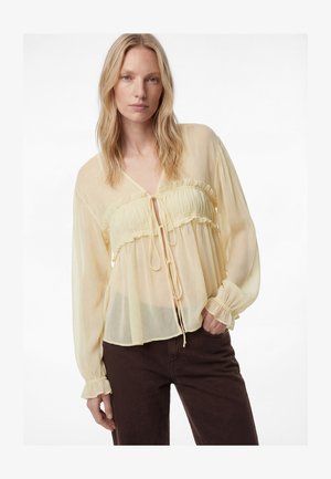 Mujer rubia con blusa translúcida de manga larga en amarillo pálido, con volantes y lazos, combinada con pantalones marrón oscuro sobre un fondo blanco.