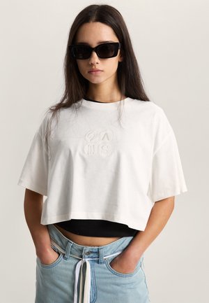 T-shirt bianca tagliata con collo rotondo, con logo in rilievo. Abbinata a jeans a vita alta azzurri e una maglia nera sottostante.