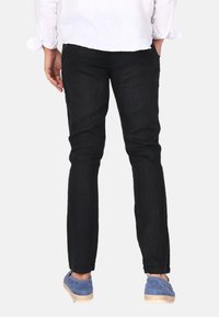 Schwarze Denim-Hosen, Slim Fit, mit glatter Textur und minimaler Naht. Kombiniert mit blauen Slippern für einen lässigen Look.