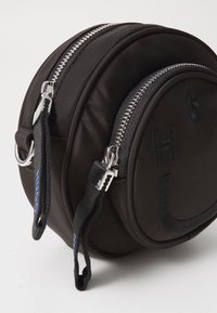 Okrągna czarna torba wykonana z gładkiego materiału, z podwójnymi srebrnymi zamkami, haftowanymi literami i krótkimi materiałowymi uchwytami z niebieskimi akcentami.