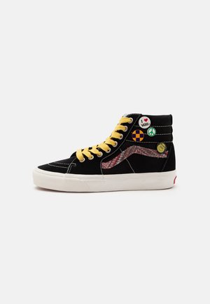 Basket montante noire avec des lacets jaunes, une semelle blanche, quatre pins ronds colorés et une bande latérale rouge et blanche à motifs.