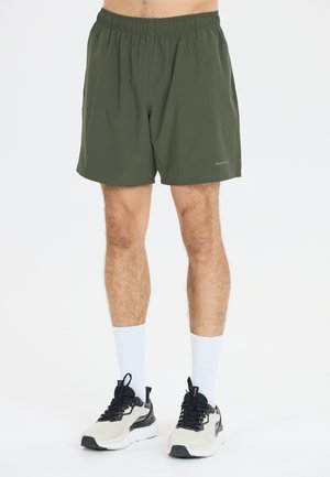Shorts vert olive avec une taille élastique, fabriqués à partir d'un matériau léger, comportant un détail de logo et une longueur mi-cuisse. Assortis à des baskets noires et blanches.