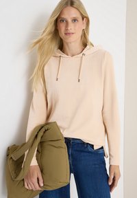 Une femme blonde portant un hoodie beige et un jean bleu tient une doudoune vert olive devant un fond neutre.