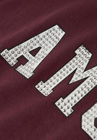 Tissu bordeaux avec de grandes lettres argentées "A" et "M" embellies de clous réfléchissants, offrant une texture lisse et un design graphique audacieux.