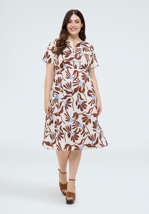 Mujer sonriente con vestido blanco hasta la rodilla con estampado floral marrón y azul y sandalias marrones de tacón alto, de pie sobre fondo blanco.