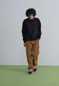 Sweatshirt negru, pantaloni cargo maro, încălțăminte sport albă și neagră. Modelul stă pe un fundal verde, împotriva unui perete simplu, surprinzând o ținută casual.
