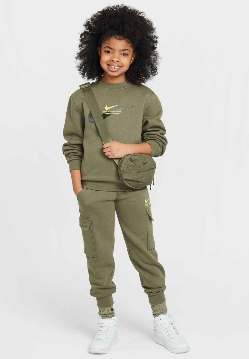Oliwkowy sweatshirt Nike z żółtym logo, zestawiony z pasującymi spodniami dresowymi w stylu cargo. Białe sneakersy, mała torebka na biodro i kręcone włosy.