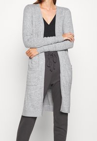 Vaaleanharmaa pitkä neuletakki, jossa avoin etuosasuunnittelu, resorireunaiset hihat ja kaksi etutaskua, valmistettu pehmeästä ja teksturoidusta neulekankaasta.