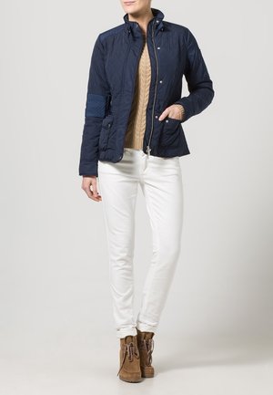 Chaqueta acolchada azul marino con cuello de pie, dos bolsillos delanteros y puños acanalados. Se lleva sobre un suéter de punto trenzado color beige y pantalones blancos, combinada con botas marrones.