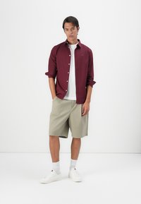 Bordeaux overhemden met knopen, gedragen over een wit T-shirt, gecombineerd met kaki shorts en witte sneakers, staande op een effen achtergrond.