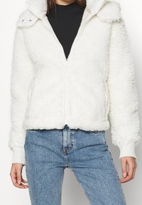Bílá kožená bunda s kožešinou na zip a kapucí, s žebrovanými manžetami a lemem, a texturovaným povrchem. V kombinaci s modrými džíny s vysokým pasem.