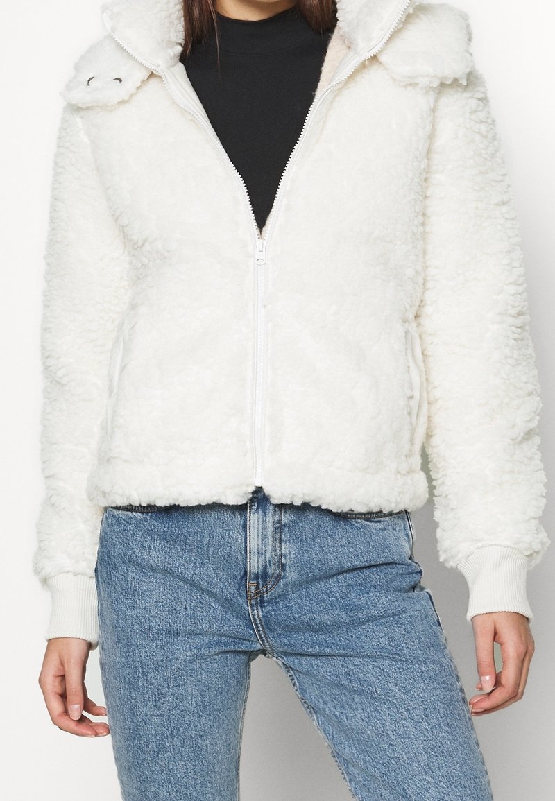 Bílá kožená bunda s kožešinou na zip a kapucí, s žebrovanými manžetami a lemem, a texturovaným povrchem. V kombinaci s modrými džíny s vysokým pasem.