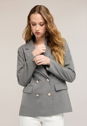 Ung kvinde med langt bølget hår justerer manchet på grå dobbeltradet blazer med nålestriber, iført store guldbelagte øreringe og hvide bukser.