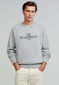 Gray sweatshirt with navy embroidered text "THE SCALPERS CO. ATELIER," featuring a round neck and ribbed cuffs, worn with beige pants.