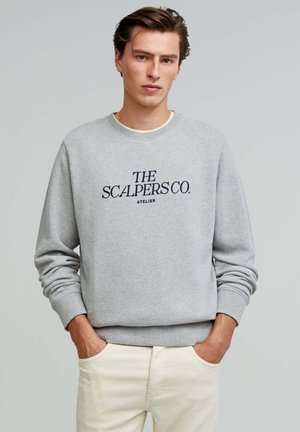 Gray sweatshirt with navy embroidered text "THE SCALPERS CO. ATELIER," featuring a round neck and ribbed cuffs, worn with beige pants.