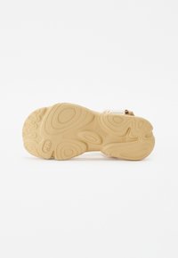 Tan rubber sandal sole with textured patterns, contoured grips, and a logo. White adjustable strap integrated into the design.