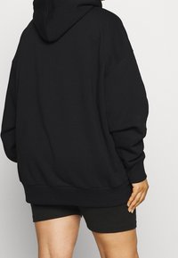 Felpa nera oversize con cappuccio regolabile, polsini e bordo a coste, realizzata in morbido tessuto testurizzato. Indossata con pantaloni corti neri.