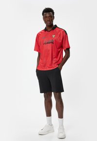 Rød atletisk trøje med sort krave og ærmer, prydet med "LEAGUE" i fed sort tekst. Bæres med sorte shorts og hvide sneakers.