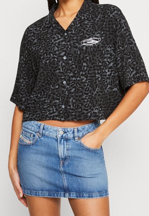 Kvinde iført en cropped, sort og grå leopardprint knap-op skjorte med en hvid oval logo, kombineret med en blå denim mininederdel.