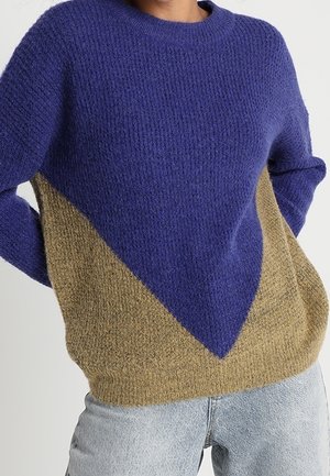 Frau trägt einen strukturierten Strickpullover mit großem dunkelblauem Dreiecksmuster auf Beige, kombiniert mit hellblauer Jeans.