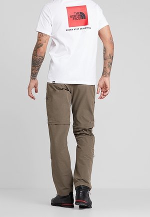 Mężczyzna z wytatuowanymi ramionami, ubrany w białą koszulkę The North Face i oliwkowe spodnie bojówki, idący do przodu na jednolitym szarym tle.