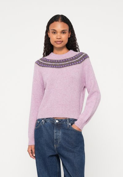 Un pull en maille violet clair avec un col rond, arborant un motif foncé sur l'empiècement. Porté avec un jean bleu taille haute et des poignets côtelés.