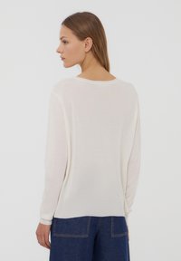 Maglione leggero in crema con maniche lunghe, scollo rotondo e texture liscia. Indossato sopra pantaloni in denim a gamba larga blu.
