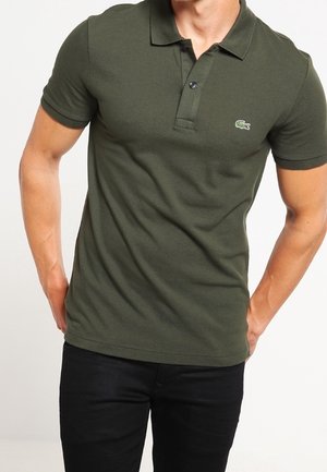 Mand iført mørkegrøn kortærmet poloshirt med to knapper, kombineret med sorte bukser, hænderne hvilende bag ryggen.