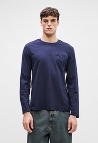 Mornarska majica z dolgimi rokavi iz bombaža, s krožnim izrezom in vezenim logotipom na levem prsnem delu. Gladka tekstura, sproščen kroj.