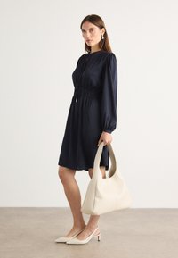 Navy dress with long sleeves, cinched waist; model holds a large cream handbag. Pointed white heels, minimalistic design, smooth texture.