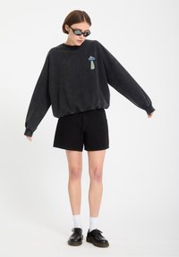 Joven con sudadera negra de gran tamaño con un parche de un OVNI y un cactus, pantalones cortos negros, calcetines blancos, zapatos negros y gafas de sol, posando.