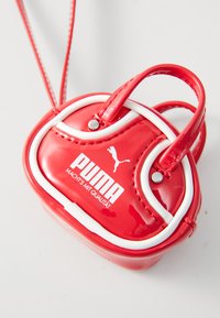 Majhna sijajna rdeča Puma torbica z belim robom, dvojni ročaji in naramnico, označena z logotipom Puma in nemškim besedilom "Macht's mit Qualität."