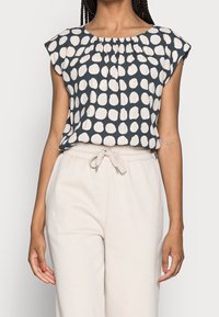 Blusa a maniche corte di colore blu navy con grandi pois bianchi, caratterizzata da un colletto plissettato, abbinata a pantaloni beige con vita regolabile.
