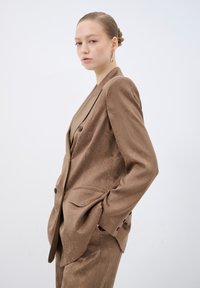 Blazer marrone con texture e vestibilità su misura, dotato di due bottoni, tasche a patta e una fodera interna a contrasto, abbinato a un outfit coordinato.