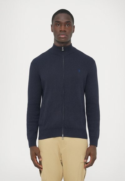 Marineblå ribstrikket cardigan med lynlås og høj krave, med sidelommer og et diskret logo på venstre bryst. Blød tekstur og tætsiddende design.