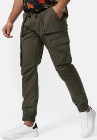 Cargo shorts i olivgrön tyg, med flera fickor, elastiska muddar och snörliv. Bärs med vita sneakers.