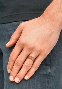 Metalowy pierścień o matowym brązowym wykończeniu, w kształcie zaokrąglonym, noszony na dłoni opierającej się na ciemnoniebieskich dżinsach. Widoczna tekstura skóry na palcach.