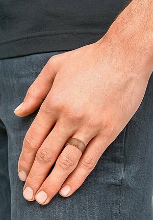 Anillo de metal con un acabado mate en color marrón, forma redondeada, llevado en una mano que descansa sobre un pantalón de mezclilla oscuro. Textura de la piel visible en los dedos.