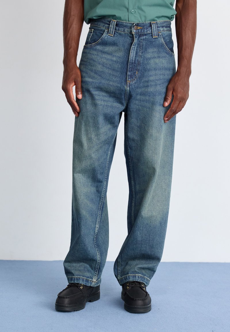 Široke jeans hlače iz modrega denima s posvetljeno obdelavo, z gumbom na zapiranju in petimi žepi, v kombinaciji s črnimi čevlji.