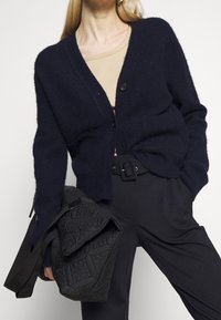 Marinblå ullcardigan med V-ringning, fyra knappar och ribbade ärmar, bärs över en beige topp, i kombination med mörka byxor och en strukturerad svart väska.