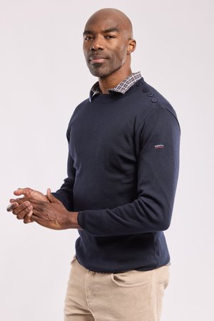 Man met gevouwen handen die een marineblauwe trui draagt over een geruit hemd en beige broek, staand tegen een effen lichte achtergrond.