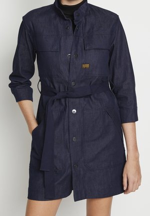 Vrouw draagt een donkerblauwe denim jurk met ceintuur, voorzakken en knoopsluitingen, de mouwen zijn opgerold tot aan de onderarmen.