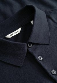 Sötét tengerészkék pólóing, puha kötött anyagból készült. Klasszikus gallérral rendelkezik, és három szürke gomb található a nyakán, egy szőtt címkével.