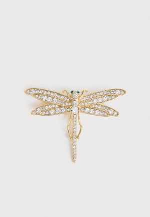 Καρφίτσα σε σχήμα χρυσής λιβελούλας διακοσμημένη με διαυγή κρύσταλλα στα φτερά και το σώμα, με δύο μικρά πράσινα πολύτιμα μάτια, σε λευκό φόντο.