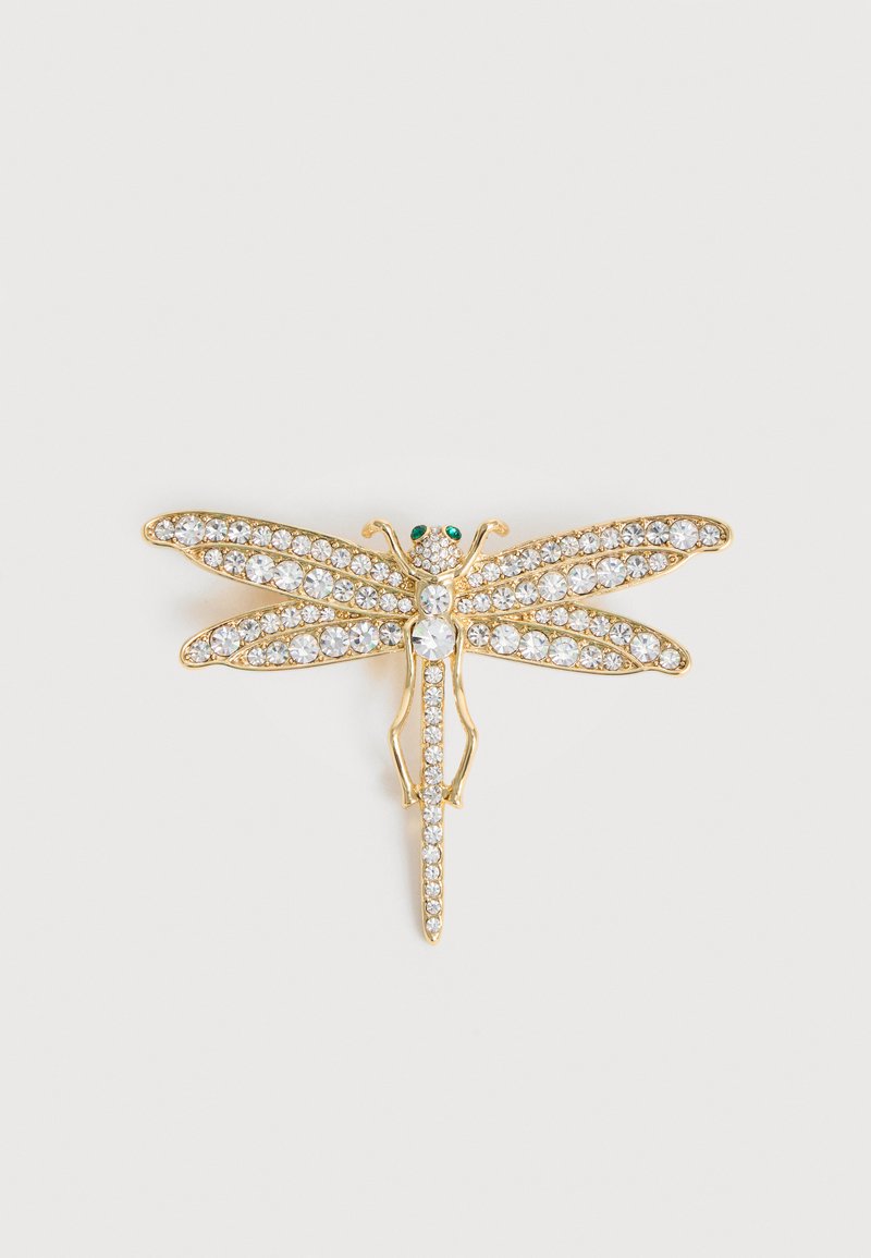 Καρφίτσα σε σχήμα χρυσής λιβελούλας διακοσμημένη με διαυγή κρύσταλλα στα φτερά και το σώμα, με δύο μικρά πράσινα πολύτιμα μάτια, σε λευκό φόντο.