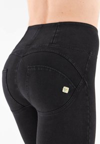 Lähikuva henkilöstä, joka käyttää korkeavyötäröisiä mustia housuja, joissa on kaarevat saumat ja pieni neliönmuotoinen metallilogo takataskussa.