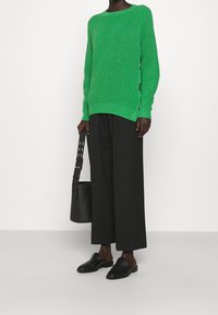 Grüner gerippter Strickpullover mit Rundhalsausschnitt und seitlichen Knopfdetails; kombiniert mit weiten schwarzen Hosen und schwarzen Slippern.